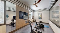 Located at 1851 Encantado Way in Kissimmee, Florida, this dedicated cycling and spin studio offers a focused fitness environment designed for high-energy indoor cycling workouts. The single-room space is outfitted with multiple Echelon smart stationary bikes, each paired with its own interactive display screen for guided on-demand or live cycling classes. A large flat-screen television is mounted on a warm wood-paneled accent wall, serving as a central focal point for group instruction or entertainment during sessions. Full-length mirrors line one wall, enhancing the sense of space and allowing users to monitor their form. The room features a stylish design with distinctive horizontal stripe molding along the upper walls, recessed lighting, and a ceiling fan that promotes air circulation during intense workouts. Durable, textured carpet tile flooring provides comfortable traction underfoot. A floor-to-ceiling glass partition wall separates the studio from an adjacent corridor, maintaining visual openness while defining the workout zone. Fitness accessories including stability balls, resistance bands, and exercise mats are also available within the space. A water fountain is conveniently accessible just outside the studio entrance, rounding out this well-equipped and thoughtfully designed cycling facility.