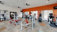 This fitness center is a thoroughly equipped, single-floor facility designed to support a wide range of workout routines. The space is characterized by bold terracotta-red accent walls paired with crisp white ceilings, creating an energizing atmosphere conducive to exercise. Durable, patterned carpet tiles cover the floor throughout, providing both comfort and traction underfoot.

The cardio zone features multiple Life Fitness treadmills equipped with personal display screens, alongside elliptical trainers and stationary bikes, all positioned to face large windows fitted with horizontal blinds. These windows allow generous natural light to fill the space while offering views of the surrounding neighborhood. Ceiling fans are installed throughout to promote airflow and comfort during workouts.

The strength training area is generously stocked with a variety of cable machines, resistance equipment, and weight benches with coordinating red upholstery. A full dumbbell rack lines one wall, accompanied by a large floor-to-ceiling mirror that visually expands the space and allows for proper form monitoring. A wall-mounted television is also present for entertainment. Recessed lighting supplements the natural light, ensuring the space remains bright and functional at all hours. The facility is accessible via glass-panel entry doors and is well-organized for an efficient, full-body workout experience.
