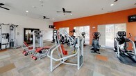 This fitness center is a thoroughly equipped, single-floor facility designed to support a wide range of workout routines. The space is characterized by bold terracotta-red accent walls paired with crisp white ceilings, creating an energizing atmosphere conducive to exercise. Durable, patterned carpet tiles cover the floor throughout, providing both comfort and traction underfoot.

The cardio zone features multiple Life Fitness treadmills equipped with personal display screens, alongside elliptical trainers and stationary bikes, all positioned to face large windows fitted with horizontal blinds. These windows allow generous natural light to fill the space while offering views of the surrounding neighborhood. Ceiling fans are installed throughout to promote airflow and comfort during workouts.

The strength training area is generously stocked with a variety of cable machines, resistance equipment, and weight benches with coordinating red upholstery. A full dumbbell rack lines one wall, accompanied by a large floor-to-ceiling mirror that visually expands the space and allows for proper form monitoring. A wall-mounted television is also present for entertainment. Recessed lighting supplements the natural light, ensuring the space remains bright and functional at all hours. The facility is accessible via glass-panel entry doors and is well-organized for an efficient, full-body workout experience.