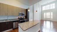This single-floor residence showcases a thoughtfully designed open layout with dramatically high ceilings that lend an airy, expansive feel throughout. The kitchen is a standout feature, equipped with stainless steel appliances including a refrigerator, range, microwave, and dishwasher. Two-toned cabinetry in warm wood and dark espresso tones frames granite countertops, which continue onto a generous peninsula island — ideal for casual dining or meal prep. Cylindrical pendant lights hang above, adding a modern, polished touch. Rich hardwood flooring extends through the kitchen and living area, where soaring walls and recessed track lighting enhance the sense of volume and openness.

The bedrooms feature plush carpeting and impressively tall ceilings, with large windows framing outdoor greenery and filling each room with natural light. Closet access is conveniently integrated. Two bathrooms offer granite-topped dark vanities, tiled tub-shower combinations with handheld fixtures, and a mix of hardwood and tile flooring. The primary bath includes an extended vanity with ample storage and connects directly to the bedroom. Throughout, the design balances contemporary finishes with a clean, neutral palette, creating a cohesive and inviting atmosphere.