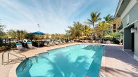 This inviting outdoor amenity space centers around a generously sized resort-style swimming pool with crystal-clear blue water, designed to offer a relaxing and recreational retreat. The pool features a freeform shape that adds a leisurely, resort-inspired character to the setting. Surrounding the pool is a well-appointed sundeck finished in warm-toned pavers, furnished with rows of cushioned lounge chairs and a variety of brightly colored market umbrellas in shades of blue, yellow, and green, providing ample shaded seating for residents and guests. The landscape is enriched by mature palm trees and lush tropical greenery, creating a vibrant, Florida-inspired atmosphere. A perimeter fence encloses the pool area, ensuring privacy and security, while the open sky above allows for abundant natural light throughout the day. The overall ambiance is bright, welcoming, and leisure-focused, offering a strong sense of outdoor living and relaxation. Note: Several of the provided interior images were captured in very low-light conditions, making interior room details indiscernible; the description reflects only what is clearly visible from the available imagery.