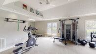 Located at 3220 Watchman Dr in Montgomery, AL, this fitness center is a thoughtfully designed single-floor facility offering a comprehensive workout environment. The space is organized into distinct functional zones, beginning with a welcoming entry foyer that features neutral gray walls, crown molding, carpeted tile flooring, and framed artwork, setting a clean and polished tone upon arrival.

The main exercise area is generously proportioned, accommodating a wide range of fitness equipment without feeling crowded. Cardio equipment includes an elliptical trainer and treadmill, positioned alongside a wall-mounted pull-up bar and a full-length mirror that enhances the sense of space. A multi-station cable machine serves as a focal point in the strength training section, complemented by a flat utility bench, a dedicated dumbbell and kettlebell rack, and medicine balls for functional training. Resistance bands and additional accessories are neatly organized on wall-mounted holders.

Vaulted ceilings in portions of the workout area add visual openness, while multiple windows along the perimeter allow abundant natural light to filter in. A ceiling fan provides added air circulation. The carpeted tile flooring throughout offers both comfort and durability, making this fitness center a practical and inviting space for a variety of training needs.
