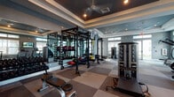 Located at 1450 Neo Square Blvd in Kissimmee, Florida, this fitness center is a thoughtfully designed, single-floor facility offering a comprehensive workout environment. The space is defined by its contemporary aesthetic, featuring cool gray walls, checkered rubber flooring in neutral tones, and tray ceilings with recessed lighting that lend a polished, upscale feel throughout.

The main workout area is generously proportioned and anchored by a large multi-functional power rack system, complete with an extensive selection of kettlebells, dumbbells, and medicine balls neatly organized on dedicated racks along mirrored walls. The mirrors amplify the sense of space and allow for proper form monitoring during training. Adjustable weight benches and a cable machine round out the strength training offerings.

A dedicated cardio zone features a row of modern treadmills positioned beneath large windows, providing natural light and exterior views that enhance the exercise experience. Wall-mounted flat-screen televisions are located throughout the space, offering entertainment across multiple workout zones.

The facility benefits from abundant natural light through large windows and a full-glass entry door, which opens to the surrounding community grounds. Ceiling fans ensure comfortable airflow during intense workouts. The space connects seamlessly to an adjacent community lounge area via glass-panel double doors, creating a cohesive and accessible amenity environment.