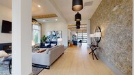 This thoughtfully designed single-floor clubhouse offers a seamless blend of modern aesthetics and functional communal spaces. Light wood-look flooring flows throughout, unifying each area while soaring ceilings with exposed wood beam accents lend an airy, architectural warmth to the interior.

The main lounge is anchored by a sleek fireplace set against a white brick surround, flanked by oversized windows that flood the space with natural light and offer views of the surrounding greenery. A statement ring chandelier and bold blue velvet seating create an inviting focal point. Adjacent to this is a second intimate seating nook featuring a sculptural wire pendant light, a mounted flat-screen TV, and coordinated lounge chairs arranged for casual gathering.

A textured stone accent wall adds dimension near the entry corridor, where a series of black pendant lights guide visitors through the expansive open layout. A dedicated game and activity area includes an arcade-style table, leather ottoman stools, and curated ocean-inspired artwork on crisp white walls.

The wet bar and dining area is outfitted with a large communal table surrounded by plush navy chairs, pendant lighting overhead, and a mounted TV framed by the same stone wall treatment. A built-in sink and open shelving complete the space. Throughout, carefully selected artwork and lush indoor plants enhance the polished yet welcoming ambiance of this well-appointed community space.