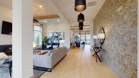 This thoughtfully designed single-floor clubhouse offers a seamless blend of modern aesthetics and functional communal spaces. Light wood-look flooring flows throughout, unifying each area while soaring ceilings with exposed wood beam accents lend an airy, architectural warmth to the interior.

The main lounge is anchored by a sleek fireplace set against a white brick surround, flanked by oversized windows that flood the space with natural light and offer views of the surrounding greenery. A statement ring chandelier and bold blue velvet seating create an inviting focal point. Adjacent to this is a second intimate seating nook featuring a sculptural wire pendant light, a mounted flat-screen TV, and coordinated lounge chairs arranged for casual gathering.

A textured stone accent wall adds dimension near the entry corridor, where a series of black pendant lights guide visitors through the expansive open layout. A dedicated game and activity area includes an arcade-style table, leather ottoman stools, and curated ocean-inspired artwork on crisp white walls.

The wet bar and dining area is outfitted with a large communal table surrounded by plush navy chairs, pendant lighting overhead, and a mounted TV framed by the same stone wall treatment. A built-in sink and open shelving complete the space. Throughout, carefully selected artwork and lush indoor plants enhance the polished yet welcoming ambiance of this well-appointed community space.