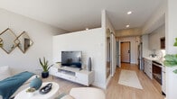 This single-floor residence showcases a contemporary design aesthetic with a cohesive palette of whites, soft grays, and warm wood-tone flooring that runs seamlessly throughout the space. The open-concept living and dining area is a standout feature, anchored by large street-facing windows that flood the room with natural light and offer views of the surrounding neighborhood. The living area comfortably accommodates both a seating arrangement and a dedicated dining space, giving the area a well-defined yet flowing layout.

A dedicated media wall with built-in storage anchors one end of the living space, while a corridor lined with arched mirrors leads toward the entry, adding visual depth. The galley-style kitchen is efficiently designed with flat-front white cabinetry, stainless steel appliances including a refrigerator and microwave, an undermount sink, and a smooth countertop workspace. Exposed concrete ceiling detail above the kitchen adds an industrial accent.

The bedroom is a calm, intimate retreat with soft neutral tones and warm lighting. The full bathroom features a tub-shower combination, a floating gray vanity with an integrated sink, and clean white finishes throughout, maintaining the unit's modern sensibility.