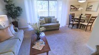 This single-floor residential space offers a comfortable and well-appointed layout designed for easy everyday living. The living area serves as the heart of the home, centered around plush wall-to-wall carpeting that adds warmth underfoot. A large sliding glass door dominates one wall, framed by elegant sheer white curtains that diffuse abundant natural light throughout the room while offering serene views of the tree-lined outdoor surroundings and a private balcony or patio beyond. The living area is anchored by a classic loveseat with decorative accent pillows, complemented by a wooden coffee table that adds a touch of warmth and character to the space. Seamlessly connected to the living area is a dedicated dining space, providing a natural flow between the two zones. The dining area features a dark wood table and chairs, ideal for everyday meals or casual entertaining. A framed cityscape artwork accents the dining wall, adding a subtle urban touch to the otherwise nature-inspired atmosphere. Together, the open layout, soft neutral tones, and generous natural light create an inviting and functional living environment that balances comfort with style.