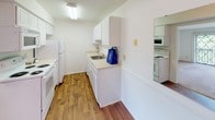This single-floor residential unit offers a clean, functional layout designed for comfortable everyday living. The galley-style kitchen is efficiently organized, featuring white cabinetry, light countertops, a stainless steel sink, and white appliances including a refrigerator, electric range, and built-in microwave. Warm hardwood-style flooring runs throughout the kitchen and connecting areas, adding a welcoming contrast to the bright white walls and cabinetry.

The living area is generously proportioned, finished with soft neutral carpeting and crisp white walls that create a bright, open atmosphere. A sliding glass door fitted with vertical blinds allows natural light to fill the space and provides access to the outdoors. The room offers ample flexibility for a variety of furniture arrangements.

The bedroom continues the neutral carpet and light-filled aesthetic, with a large window offering views of surrounding greenery. A mirrored sliding-door closet enhances the sense of space while providing practical storage.

A dedicated laundry nook houses a stacked front-loading washer and dryer alongside white upper cabinetry and a built-in countertop workspace. The full bathroom is neatly appointed with a vanity sink, a large mirror, and a shower-tub combination, all finished in a clean, neutral palette with rich hardwood-style flooring.
