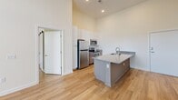 Located at 1601 Overbrook Rd in Richmond, Virginia, this single-floor residence offers a clean, contemporary design with an emphasis on open living and modern finishes throughout. The space is characterized by soaring ceilings that lend an airy, expansive feel to every room, complemented by smooth, neutral-toned walls and light wood-style flooring that flows seamlessly from room to room.

The kitchen serves as a central focal point, anchored by a substantial granite-topped island with an integrated stainless steel sink. White upper cabinetry extends toward the tall ceilings, providing generous storage, while darker lower cabinets add a stylish contrast. A full suite of stainless steel appliances, including a refrigerator, range, and microwave, completes the well-equipped cooking space. A subway tile backsplash adds a refined, contemporary touch.

The living and bedroom areas are bright and open, benefiting from large multi-pane windows that draw in abundant natural light and offer views of the surrounding neighborhood. The tall ceilings further enhance the sense of space throughout these rooms.

The bathroom is thoughtfully finished with a granite-topped vanity, warm-toned cabinetry, a large mirror, and stone-look tile flooring. A full tub-shower combination with brushed nickel fixtures provides both comfort and functionality. Overall, this unit presents a polished, move-in-ready living environment with consistent, high-quality finishes.