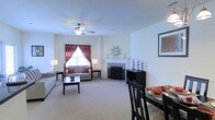 This single-floor residence showcases an open-concept layout that connects the living and dining areas in a cohesive, functional arrangement. The living room is anchored by a tiled corner fireplace, which serves as a natural focal point, accented by a decorative sunburst mirror mounted above the mantle. Neutral-toned carpet throughout the space provides a warm, comfortable feel underfoot. A ceiling fan with light fixture is centrally positioned overhead, ensuring both air circulation and ambient lighting. A window dressed with rich burgundy curtains allows natural light to filter into the living area, complementing the light, neutral wall tones. The living room comfortably accommodates a sofa and loveseat arrangement alongside a rectangular coffee table. The dining area, situated adjacent to the living space, features a dark-finished table and chair set, adding a bold contrast to the lighter surroundings. Framed wall art adds a finishing decorative touch. The overall ambiance is welcoming and well-balanced, blending functional furnishings with tasteful decorative elements for comfortable everyday living.