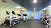 This single-floor fitness center offers a functional and well-organized workout space designed for cardio-focused exercise. The room features rubber flooring that provides durability and shock absorption, making it well-suited for high-impact activity. A row of cardio machines — including treadmills and an elliptical trainer — lines the space, offering users a variety of aerobic workout options. Overhead, flush-mounted and recessed lighting illuminates the room evenly, complemented by a dark-painted ceiling that lends a modern, defined aesthetic to the interior. A glass partition wall adds a sense of openness while visually separating distinct areas within the facility. Ventilation is provided through ceiling vents, ensuring comfort during exercise. A standard door provides access to an adjoining room or corridor, suggesting the fitness center may be part of a larger building amenity suite. The overall design is clean, practical, and purpose-built, prioritizing utility and ease of use for residents or occupants seeking a convenient on-site exercise option.