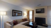 This single-floor residence offers a thoughtfully arranged layout connecting its main living spaces in a cohesive flow. The living room serves as a central gathering area, featuring a textured ceiling, a classic fireplace with a white mantel, and a window dressed in warm golden drapes that allow natural light to filter through. A wall-mounted flat-screen television and framed landscape artwork add to the room's polished character. The space accommodates a generously sized sectional sofa, anchored by a dark wood coffee table, creating a welcoming and functional seating arrangement.

Adjacent to the living area, the dining space features soft carpeting, track lighting overhead, and a round dark wood dining table surrounded by upholstered cream chairs. Framed artwork lines the hallway wall, adding visual interest to the transition between rooms. The kitchen, visible from the dining area, includes wooden cabinetry, a stainless steel refrigerator, and a breakfast bar, providing practical functionality. Throughout the residence, a neutral color palette of warm whites and beiges creates a calm, cohesive ambiance well-suited for comfortable everyday living.