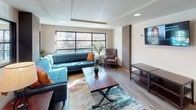 Located at 95 Burnett Ave S in Renton, Washington, this single-floor community space combines modern design with a warm, inviting atmosphere. The layout flows seamlessly from a well-lit corridor entrance — accented by a bold teal feature wall, wood-look flooring, and recessed ceiling lighting — into an open lounge area. The lounge is generously proportioned, anchored by a tufted teal sectional sofa, a rustic wood coffee table, and a patterned area rug that adds a pop of color. Large multi-pane windows flood the space with natural light and offer views of the surrounding greenery, while tray ceilings and recessed lighting contribute to a polished architectural feel. A wall-mounted mini-split unit ensures year-round climate comfort. Adjacent to the lounge, a kitchenette provides practical amenities including warm wood cabinetry, dark granite tile countertops, a double stainless steel sink, and track lighting overhead — making the space well-suited for gatherings and events. Throughout the space, decorative artwork, indoor plants, and thoughtful furnishings create a cohesive, community-oriented environment. The combination of contemporary finishes, durable flooring, and an open, connected layout makes this a functional and welcoming shared space.