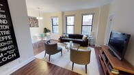 This thoughtfully designed urban residence offers a seamless blend of comfort and contemporary style across a single level. The open-plan living and dining area serves as the heart of the home, featuring rich hardwood flooring, warm neutral walls, and crown molding that lends an elegant touch. Multiple black-framed windows flood the space with natural light while framing views of the surrounding cityscape. The living area is anchored by a dark tufted sofa, paired with rounded accent chairs and a glass coffee table, all centered on a plush area rug. A wooden media console with a flat-screen television completes the arrangement. The adjoining dining area accommodates a glass-top round table with white chairs, lit by a classic chandelier overhead. The kitchen is outfitted with dark cabinetry, stainless steel appliances including a refrigerator, microwave, and electric range, and clean white countertops. The bedroom offers a tranquil retreat with plush carpeting, crown molding, ample storage via a large multi-drawer dresser, and a window with city views. The bathroom features a dark vanity with a white countertop, a mirrored medicine cabinet, and clean white tile work throughout, delivering a crisp, functional finish.