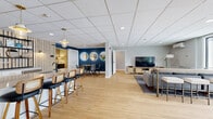 Located at 155 Gleason Lake Rd in Wayzata, Minnesota, this single-floor amenity space offers a thoughtfully designed layout that balances communal living, fitness, and productivity. The open common area serves as the heart of the space, featuring light hardwood flooring, a drop tile ceiling with recessed lighting, and a warm, contemporary aesthetic. A bar-height counter with cane-back stools creates a casual dining and gathering zone, while a bold navy accent wall adorned with three round backlit mirrors defines an intimate dining nook. A comfortable lounge area with a sectional sofa, flat-screen TV, and media console rounds out the social space. Framed botanical artwork and lush potted plants add a refined, welcoming ambiance throughout. The exercise room is comprehensively outfitted with cardio equipment including a treadmill, stationary bike, and elliptical, alongside a full dumbbell rack, adjustable bench, and wall-mounted yoga mats. Floor-to-ceiling mirrors, rubberized flooring, a striking wood slat accent wall, and a dedicated mini-split HVAC unit make it a fully functional fitness facility. The private office features a built-in dual workstation beneath an elegant drum pendant light, accented by textured wallcovering, offering a quiet and polished workspace. Connecting hallways tie each area together seamlessly.