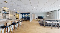 Located at 155 Gleason Lake Rd in Wayzata, Minnesota, this single-floor amenity space offers a thoughtfully designed layout that balances communal living, fitness, and productivity. The open common area serves as the heart of the space, featuring light hardwood flooring, a drop tile ceiling with recessed lighting, and a warm, contemporary aesthetic. A bar-height counter with cane-back stools creates a casual dining and gathering zone, while a bold navy accent wall adorned with three round backlit mirrors defines an intimate dining nook. A comfortable lounge area with a sectional sofa, flat-screen TV, and media console rounds out the social space. Framed botanical artwork and lush potted plants add a refined, welcoming ambiance throughout. The exercise room is comprehensively outfitted with cardio equipment including a treadmill, stationary bike, and elliptical, alongside a full dumbbell rack, adjustable bench, and wall-mounted yoga mats. Floor-to-ceiling mirrors, rubberized flooring, a striking wood slat accent wall, and a dedicated mini-split HVAC unit make it a fully functional fitness facility. The private office features a built-in dual workstation beneath an elegant drum pendant light, accented by textured wallcovering, offering a quiet and polished workspace. Connecting hallways tie each area together seamlessly.