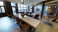 This contemporary lounge space is thoughtfully designed to blend functionality with modern aesthetics. At its center sits a striking, large-format speckled white stone communal table surrounded by cushioned swivel bar stools with patterned upholstery, making it ideal for collaborative work or casual gatherings. The open layout flows seamlessly into a comfortable seating area furnished with plush purple and earth-toned armchairs arranged around low circular coffee tables, creating an inviting atmosphere for conversation and relaxation. Warm wood paneling lines the walls, adding depth and a refined texture to the interior. A flat-screen television is mounted flush against a feature wall, enhancing the lounge's versatility. Floor-to-ceiling windows along the perimeter flood the space with abundant natural light and provide expansive views of the surrounding urban environment, while sheer curtains offer adjustable privacy. Overhead, a combination of recessed lighting and distinctive gold globe pendant fixtures cast a warm, ambient glow throughout. The flooring features neutral-toned wood-look planks that tie together the earthy and rich color palette of the furnishings. Overall, the space exudes a sophisticated, welcoming character suited for both productive and social use.
