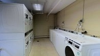 This single-floor laundry room is designed for practical, high-capacity use. The space features a row of freestanding top-load washing machines and dryers lined along one wall, complemented by a bank of stacked washer-dryer units along the opposite side, maximizing the available floor space. The room is finished with light-toned tile flooring, which is easy to maintain and clean. Overhead, exposed piping and ductwork run along the ceiling, contributing to the room's utilitarian character, while a fluorescent light fixture provides adequate illumination throughout. The walls are finished in a neutral tone, keeping the environment straightforward and functional. A baseboard heating unit runs along the base of one wall, ensuring the space remains comfortable year-round. The layout is efficiently organized, allowing users to move between machines with ease. Overall, this laundry room prioritizes functionality and capacity, making it well-suited for shared or communal use within a residential or commercial building.