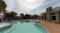 This single-story amenity facility delivers a well-rounded recreational experience through two distinct areas: a fully equipped fitness center and a resort-style outdoor pool. The fitness center features wood-look plank flooring and is lined with large windows along one wall, allowing generous natural light to fill the space and providing views of the surrounding greenery. Full-length wall mirrors visually expand the room while ceiling fans maintain comfortable airflow. The equipment selection is comprehensive, including treadmills, elliptical machines, a multi-station cable machine, weight benches, a full rack of hex dumbbells, and a vertical medicine ball storage tower stocked with varied weights. The layout accommodates both cardio and strength training routines efficiently within the open floor plan. Outside, the freeform swimming pool is a standout feature, with a curving design and clear turquoise water. A dedicated pool deck surrounds the area and is furnished with blue lounge chairs and a shaded umbrella station. Stone-accented architectural elements on the adjacent building complement the tropical landscaping of mature palm trees and manicured hedges, creating a relaxed, resort-like atmosphere beneath an open sky.