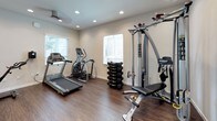 Located at 912 3rd Avenue in Chula Vista, California, this fitness center offers a comprehensive workout facility spread across a single floor. The space is designed with a clean, modern aesthetic, featuring neutral-toned walls, crisp white trim, and durable wood-style flooring throughout that gives the gym a polished, professional feel.

The facility is divided into dedicated workout zones, each serving a distinct fitness purpose. One area houses cardio equipment, including a treadmill and an elliptical machine, positioned beneath large windows fitted with white blinds that allow natural light to fill the space. Adjacent to the cardio zone is a strength training area equipped with a Hoist cable machine system and a full rack of dumbbells, offering a wide range of resistance options for all fitness levels.

A separate functional training room rounds out the facility, stocked with stability balls, medicine balls, resistance bands, suspension trainers, foam rollers, and a spin bike — ideal for group exercise or cross-training routines. Recessed lighting throughout ensures consistent illumination across all areas, while ceiling fans provide added comfort during intense workouts. Additional amenities include wall-mounted equipment dispensers and clearly posted instructional signage. The overall ambiance is bright, organized, and purpose-built for an effective fitness experience.