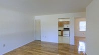 This single-floor space showcases a clean, open layout highlighted by warm hardwood flooring that runs throughout the main living area, reflecting light and contributing to a bright, welcoming ambiance. The living area is generously proportioned, with white walls and high ceilings that enhance the sense of openness. A bifold-door closet is conveniently integrated into the main living space, offering practical storage. An open passthrough connects the living area to the kitchen, which features medium-toned wood cabinetry providing ample storage, a white gas range, and a dishwasher. The kitchen's layout is functional and compact, making efficient use of the available space. The overall design is straightforward and neutral, with white walls throughout that provide a versatile backdrop. Natural light appears to filter into the space, further enhancing its airy feel. The combination of hardwood floors, classic cabinetry, and an open-concept connection between the living and kitchen areas gives this space a practical yet comfortable character suited to everyday living.