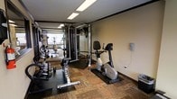 This fitness center offers a practical and functional space designed to meet a wide range of exercise needs. The room features a combination of carpet and rubber mat flooring, providing comfort and safety across different workout zones. A full rack of dumbbells lines one side of the space, accompanied by a dedicated free weights area ideal for strength training. A multi-station cable machine anchors the center of the room, offering versatile resistance training options. Cardio equipment, including an upright stationary bike, is positioned along the walls for convenient use. Large mirrors span a significant portion of the walls, enhancing the sense of space while allowing users to monitor their form during exercise. Overhead fluorescent lighting ensures the entire area is well-lit throughout the day. The neutral-toned walls contribute to a clean and focused atmosphere. The layout is thoughtfully organized to maximize usable floor space, allowing for comfortable movement between equipment. Overall, this fitness center provides a comprehensive and accessible environment suited for both cardio and strength-based training routines.