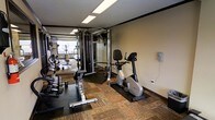 This fitness center offers a practical and functional space designed to meet a wide range of exercise needs. The room features a combination of carpet and rubber mat flooring, providing comfort and safety across different workout zones. A full rack of dumbbells lines one side of the space, accompanied by a dedicated free weights area ideal for strength training. A multi-station cable machine anchors the center of the room, offering versatile resistance training options. Cardio equipment, including an upright stationary bike, is positioned along the walls for convenient use. Large mirrors span a significant portion of the walls, enhancing the sense of space while allowing users to monitor their form during exercise. Overhead fluorescent lighting ensures the entire area is well-lit throughout the day. The neutral-toned walls contribute to a clean and focused atmosphere. The layout is thoughtfully organized to maximize usable floor space, allowing for comfortable movement between equipment. Overall, this fitness center provides a comprehensive and accessible environment suited for both cardio and strength-based training routines.