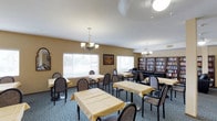 The Community Room at 2525 Highland Ave in National City is a thoughtfully designed shared space built to foster connection and comfort among its residents and visitors. The room is finished with warm, tan-toned walls that create an inviting and relaxed atmosphere throughout. Elegant chandelier pendant lights hang from the white ceiling, casting a warm glow over the dining area, which is furnished with multiple round tables dressed in yellow tablecloths and surrounded by cushioned chairs — ideal for communal meals or social gatherings. The patterned blue carpet adds a decorative touch underfoot, complementing the overall color palette of the room. Large windows fitted with white horizontal blinds line one wall, allowing natural light to filter into the space while maintaining privacy. On the opposite side of the room, a substantial dark wood bookcase spans floor to near-ceiling, filled with an extensive collection of books and media, offering residents a convenient library resource. A cozy seating area is positioned near the bookcase, providing a quiet nook for reading or relaxed conversation. Overall, the Community Room is a versatile, well-appointed space designed to serve both social dining and leisurely activities.