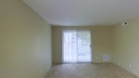 This single-floor residential space presents a clean, functional layout designed for comfortable everyday living. The living area is the focal point of the space, characterized by smooth, light-colored walls in a warm neutral tone that create a calm and welcoming atmosphere. Carpeted flooring in a complementary beige hue runs throughout, adding softness underfoot and contributing to the overall understated aesthetic.

A prominent sliding glass door serves as both a design feature and a practical element, flooding the room with natural light and providing direct access to the outdoors. Vertical blinds fitted to the door allow for flexible light control and privacy. Through the door, glimpses of outdoor greenery are visible, offering a pleasant connection to the exterior environment.

The ceiling is smooth and white, enhancing the sense of openness and airiness within the room. A wall-mounted vent is present, indicating a built-in climate control system. The overall design of the space is straightforward and versatile, with neutral finishes that provide a blank canvas easily adaptable to a variety of furnishing styles. The space is well-suited for those seeking a simple, move-in ready environment with access to natural light and the outdoors.