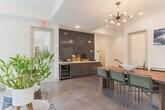 Located at 222 Peters St SW in Atlanta's dynamic Castleberry Hill neighborhood, this single-floor space offers a thoughtfully designed communal environment that blends comfort and function. The lounge area anchors the space with a rich green leather sofa accented by gold and olive throw pillows, set atop a bold geometric-patterned rug. Industrial-chic drum side tables and lush potted greenery add warmth and character, while floor-to-ceiling windows dressed with charcoal drape panels flood the space with natural light and offer street-facing views. A dedicated dining zone features a solid wood communal table surrounded by sleek metal chairs, illuminated by a striking Sputnik-style chandelier. A wall-mounted display screen and curated artwork contribute to the space's gallery-like aesthetic. The coffee bar station is a standout feature, showcasing a dramatic floor-to-ceiling black hexagonal tile backsplash, sage-green cabinetry, white quartz countertops, and a fully stocked beverage setup. Polished concrete flooring ties each zone together seamlessly, lending an urban, contemporary feel throughout. Frosted glass doors provide access to an adjacent meeting room, offering a more private setting within the overall open-plan layout.