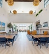 Located at 1900 E Inland Empire Blvd in Ontario, California, this single-floor community space offers a generously sized, open-concept layout spanning a substantial footprint. The design is defined by dramatically soaring ceilings with large black-framed clerestory windows that flood the interior with natural light, creating an airy and uplifting atmosphere throughout.

The entrance welcomes visitors with light wood-look flooring, oversized planters housing lush tropical trees, and floor-to-ceiling black-framed glass windows overlooking the exterior. The main gathering area transitions between elegant herringbone-patterned tile and wide-plank light wood flooring, visually delineating distinct functional zones. Seating arrangements include casual lounge groupings with cognac leather chairs flanking a fireplace, communal dining tables with navy and wood chairs, and soft seating areas anchored by landscape artwork and potted olive trees.

Built-in warm wood shelving units with integrated lighting serve as focal points, displaying curated décor alongside a wall-mounted television. Statement amber globe pendant lights hang from the high ceilings, adding warmth and sophistication. Black-framed glass double doors provide transitions between zones and lead to outdoor areas. The overall aesthetic is modern and refined, balancing bright whites with warm wood tones, greenery, and carefully selected furnishings.
