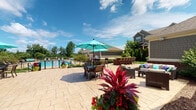 Located at 13968 John Humphrey Dr in Orland Park, Illinois, this single-level outdoor amenity space offers a well-appointed community retreat designed for relaxation and social gatherings. The centerpiece is a resort-style swimming pool enclosed by a wrought iron fence, surrounded by a concrete pool deck lined with lounge chairs, ideal for sunbathing on warm summer days. Adjacent to the pool is an expansive paver patio featuring multiple distinct seating areas. Dining tables shaded by vibrant teal umbrellas provide comfortable al fresco dining, while a dedicated lounge area with all-weather wicker furniture and colorful accent pillows creates an inviting space for casual conversation. A row of Adirondack-style chairs lines a secondary section of the patio, offering additional spots to take in the open views of the surrounding greenery. The space is accented by richly planted containers and manicured garden beds bursting with bold red tropical foliage, adding striking color and visual interest throughout. Neatly trimmed boxwood shrubs and mature trees frame the perimeter, providing natural privacy and shade. Large glass-paned doors connect the outdoor patio to an interior facility, allowing seamless indoor-outdoor flow. String lights overhead hint at the space's suitability for evening gatherings, rounding out a well-designed outdoor environment suited for community living.
