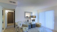 This single-story space presents a welcoming living area characterized by light, neutral walls that create a clean and airy atmosphere. The room is finished with wall-to-wall carpeting, adding warmth and comfort underfoot. Natural light filters in through a large window dressed with vertical blinds, brightening the space and enhancing its open feel. A decorative mosaic-framed mirror serves as a focal point on the main wall, complementing a glass-and-metal console table below, which is styled with vases, a table lamp, and various decorative objects. Seating includes a boldly patterned floral accent chair, a pair of upholstered ottomans, and a rounded side chair, offering a casual yet curated arrangement. A glass-top accent table is also visible, adding a contemporary touch. The ceiling features a textured finish with a visible HVAC vent, indicating a standard residential build. A hallway visible in the background suggests additional rooms beyond the living area. Overall, the space conveys a comfortable, move-in-ready aesthetic with thoughtful decorative styling throughout.
