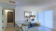 This single-story space presents a welcoming living area characterized by light, neutral walls that create a clean and airy atmosphere. The room is finished with wall-to-wall carpeting, adding warmth and comfort underfoot. Natural light filters in through a large window dressed with vertical blinds, brightening the space and enhancing its open feel. A decorative mosaic-framed mirror serves as a focal point on the main wall, complementing a glass-and-metal console table below, which is styled with vases, a table lamp, and various decorative objects. Seating includes a boldly patterned floral accent chair, a pair of upholstered ottomans, and a rounded side chair, offering a casual yet curated arrangement. A glass-top accent table is also visible, adding a contemporary touch. The ceiling features a textured finish with a visible HVAC vent, indicating a standard residential build. A hallway visible in the background suggests additional rooms beyond the living area. Overall, the space conveys a comfortable, move-in-ready aesthetic with thoughtful decorative styling throughout.