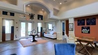 This single-floor space makes a strong first impression with its grand, open layout and refined architectural details. The main area is anchored by soaring ceilings accented with decorative crown molding and a dramatic arched entryway, lending the space a sense of grandeur and openness. Large glass doors at the entrance flood the interior with natural light, offering views of lush greenery outside.

The interior design follows a coastal-inspired aesthetic, featuring a warm wood-look flooring throughout that complements the neutral wall tones. A central seating area is styled with a crisp white sofa adorned with orange and blue accent pillows, positioned atop a coral-patterned area rug. A round decorative table with a sculptural centerpiece anchors the entryway, while an elegant hanging pendant light adds character overhead.

Architectural columns with detailed millwork define the transitional spaces, creating natural divisions between areas without sacrificing the sense of openness. A side area features additional seating with plush blue velvet chairs, suggesting a lounge or consultation zone. Deeper within, an accent wall in a rich terracotta-orange hue adds warmth and visual contrast. Informational boards and framed displays hint at a professional or community-oriented purpose, rounding out this polished and welcoming environment.