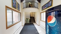 Moore Lake Apartments offers a thoughtfully designed common entry and lobby area that makes a strong first impression. The entrance is accessed through a glass-paneled door framed in warm wood trim, offering a view of lush greenery and a paved walkway outside. Security is provided via an overhead camera system, and building management information is clearly posted at the entry.

Inside, the lobby opens into a striking two-story atrium-style space with soaring vaulted ceilings supported by exposed dark beams. A curved upper-level balcony with elegant wrought-iron railings overlooks the main floor below, adding a refined architectural touch. The walls are adorned with framed artwork in rich, jewel-toned colors, lending character and warmth to the common area.

Natural light filters through large windows on both sides of the lobby, illuminating the white tile flooring accented by a central runner rug that guides residents toward the staircase. Both a staircase with handrails and an elevator are available, ensuring accessibility for all residents. The overall ambiance of the lobby is welcoming and well-maintained, reflecting a building that balances practicality with an appealing sense of community and design.
