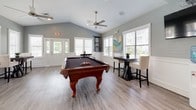 This well-appointed single-floor community space blends functionality and leisure in a bright, welcoming environment. The main gathering area is anchored by a classic wood-framed pool table set beneath a sleek ceiling fan, with vaulted ceilings that lend an open, airy feel. Light gray walls, white trim, and wood-look plank flooring run throughout, creating a cohesive and modern aesthetic. Multiple windows dressed with white horizontal blinds allow abundant natural light to fill the room, offering views of the surrounding greenery.

The kitchen is thoughtfully designed with crisp white cabinetry, rich dark granite countertops, and stainless steel appliances including a full-size refrigerator and microwave. A spacious peninsula with bar-height seating accommodates multiple guests and serves as a natural gathering point between the kitchen and the main lounge area.

Dining and seating options are plentiful, with several high-top tables paired with upholstered chairs positioned throughout the space, ideal for casual socializing or small group gatherings. The layout flows openly from the kitchen and bar area into the game and lounge space, making the room equally suited for events, recreation, or everyday use. The overall ambiance is clean, inviting, and purposefully designed for communal enjoyment.