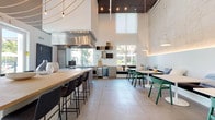 This single-floor communal space showcases a striking contemporary design defined by dramatically tall ceilings that create an airy, expansive atmosphere. The open-plan layout seamlessly integrates a fully equipped demonstration kitchen with a social dining area, making it well-suited for gatherings and culinary events.

The kitchen zone is outfitted with professional-grade stainless steel appliances, including a built-in oven, beverage cooler, and a large overhead ventilation hood. A central island with a butcher-block top provides ample prep space and doubles as a bar-height seating counter with sleek black stools. Wood-toned cabinetry and open shelving add warmth against the crisp white walls, while large windows frame views of lush outdoor greenery, filling the space with generous natural light.

The dining area features a mix of seating options, including built-in upholstered banquette seating along a raw concrete-textured wall, paired with white pedestal tables and teal accent chairs. Polished concrete floors tie the zones together, while an artistic arrangement of pendant lighting and globe string lights adds character overhead. A wall-mounted display screen and additional counter seating complete this versatile, design-forward communal environment.