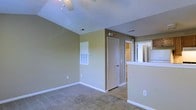 This single-floor residential space presents a practical and comfortable layout designed for everyday living. The main living area is finished with wall-to-wall carpeting that adds warmth underfoot, while neutral olive-green walls and crisp white baseboards create a clean, understated aesthetic throughout. A ceiling fan is centrally positioned overhead, providing both air circulation and ambient lighting to the space.

The textured ceiling adds a subtle architectural detail, complementing the straightforward design of the room. A large HVAC vent panel is mounted on the wall, indicating a built-in climate control system integrated into the space. Visible door access leads to an interior room or closet, suggesting additional storage or private living quarters beyond the main area.

To one side, a half-wall partition opens into the kitchen, allowing for a connected flow between the living and cooking areas while maintaining a degree of separation. The kitchen area, partially visible, appears to include appliances and cabinetry, contributing to the functional layout of the home. Overall, the space offers a straightforward, well-maintained environment with consistent finishes and a practical floor plan suited for comfortable day-to-day living.