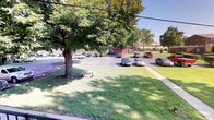 This single-story property presents a welcoming exterior defined by a generously sized, well-kept lawn that stretches across the front of the building. A large, mature shade tree anchors the green space, providing natural canopy and a shaded outdoor area complete with a small bench or picnic table positioned beneath its branches — ideal for relaxing outdoors.

The paved parking area is clearly striped and accommodates multiple vehicles with ease, suggesting a practical layout designed for convenience. Brick construction is visible in the background, indicative of durable, classic building materials that contribute to the property's sturdy and established appearance.

The surrounding landscape is lush and green, with additional mature trees lining the perimeter, enhancing privacy and the overall aesthetic of the grounds. The open sky and abundant natural light create a bright, airy atmosphere throughout the exterior. The combination of maintained greenery, ample parking, and solid brick architecture gives the property a well-organized and functional character suited to a variety of uses.
