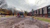 Located at 360 Broad Meadows Blvd in Columbus, Ohio, this single-story property sits within a residential community in the 43214 zip code. The most prominent feature visible is a spacious outdoor playground area, which is well-equipped with a variety of colorful recreational structures. The playground includes a multi-component play set with slides, climbing elements, and a blue-roofed tower, complemented by a standalone colorful loop-style climbing structure in the foreground. The play surface is covered with wood chip mulch, providing a safety-conscious ground cover throughout the area. Mature deciduous trees surround the perimeter, lending a sense of established greenery to the grounds, while several evergreen trees add year-round visual interest. The adjacent building features a multi-unit construction with brown siding and brick accents, consistent with a residential complex setting. Picnic benches are also visible near the playground, offering seating for residents and visitors. The overall outdoor environment is open and functional, designed to serve a community of residents, with ample space for recreation and leisure activities.