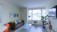 This single-floor space offers a thoughtfully designed interior that balances comfort and modern style. The living area serves as the focal point of the space, anchored by a large corner window arrangement that floods the room with abundant natural light and offers views of the surrounding greenery outside. The walls are finished in a soft, neutral green-white tone that enhances the airy feel of the room. Hardwood-style flooring runs throughout, adding warmth and continuity to the space. The living area is furnished with a rich brown leather sofa accented with colorful throw pillows in blue and green hues, paired with a sleek, modern glass and chrome coffee table atop a geometric-patterned area rug. A minimalist white accent chair with a bright cushion adds a playful pop of color near the window. Framed art prints line one wall, adding a curated, gallery-like touch to the room. A tall potted tropical plant in the corner brings a refreshing natural element indoors. The clean lines, modern furnishings, and generous natural light combine to create a bright, inviting atmosphere well-suited for both relaxation and everyday living.