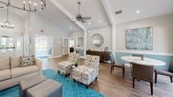 This single-story community space offers a thoughtfully designed interior that blends comfort and functionality. The main lounge area immediately impresses with soaring vaulted ceilings, recessed lighting, and a ceiling fan that enhances the open, airy atmosphere. Warm wood-look flooring runs throughout, complemented by a two-tone wall treatment featuring soft gray upper walls and a muted teal wainscoting below, creating a cohesive coastal aesthetic. Tasteful furnishings — including patterned accent chairs, a plush sofa, and a round dining table — are arranged across the space, providing both social and functional seating areas. A decorative round mirror and curated artwork add refined visual interest to the walls. Large windows and glass doors frame views of an outdoor pool area, flooding the interior with natural light. A dedicated business center room features dual workstations with computer monitors, stylish upholstered chairs, and its own teal area rug, connected to the main area via a large interior window. The kitchenette is equipped with white cabinetry, stainless steel appliances, a granite countertop breakfast bar with bar seating, and a window overlooking lush greenery. Together, these spaces form a polished, welcoming environment ideal for community gatherings and everyday use.