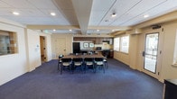 This well-appointed single-floor community space is thoughtfully designed to accommodate a variety of group activities and social gatherings. The layout comprises several interconnected zones, each serving a distinct purpose while maintaining a cohesive aesthetic throughout.

The main gathering area centers around a large rectangular dining table surrounded by dark-framed chairs, ideal for group meals or meetings. Adjacent to this is a fully equipped kitchen featuring dark wood cabinetry, a double-basin sink, a dishwasher, a microwave, and a gas range — well-suited for catering or communal cooking events.

A separate lounge area offers comfortable green accent chairs arranged around a wall-mounted flat-screen television and a dark wood credenza, creating an inviting space for casual socializing or presentations. Interior pass-through windows connect this lounge to the main gathering room, adding visual openness between the zones.

Throughout the space, warm yellow-toned walls, dark blue carpet, and recessed ceiling lighting contribute to a comfortable and functional atmosphere. Multiple windows fitted with slatted blinds allow natural light to filter in. Framed artwork adorns the walls, adding a polished finishing touch. A direct exterior door provides convenient access, rounding out this practical and welcoming multipurpose facility.