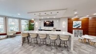 This contemporary amenity lounge in West Chester, PA is a well-appointed communal space spanning a generous single-floor layout. The design blends modern aesthetics with inviting functionality, featuring light wood-look flooring in the entry and transition areas, complemented by patterned carpet tiles throughout the main gathering zones. Structural columns are cleanly finished in white, contributing to the open, airy feel of the space.

A striking circular ring chandelier anchors the entryway, drawing the eye upward to the bright white ceiling with recessed lighting. Large windows along the perimeter flood the interior with natural light and provide views of the surrounding outdoor areas. Warm wood-paneled accent walls add contrast and depth to the otherwise neutral palette.

The lounge is organized into distinct activity zones. A prominent marble-topped bar counter with upholstered swivel stools serves as a central social hub, accompanied by a wall-mounted TV and pendant sconce lighting. A separate white high-top table offers additional seating. A billiards table anchors a dedicated game area, while a life-size Scrabble board wall display adds a playful, decorative focal point. Intimate seating nooks with modern chairs and glass accent tables round out the space, creating a versatile and engaging environment for residents.