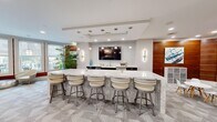 This contemporary amenity lounge in West Chester, PA is a well-appointed communal space spanning a generous single-floor layout. The design blends modern aesthetics with inviting functionality, featuring light wood-look flooring in the entry and transition areas, complemented by patterned carpet tiles throughout the main gathering zones. Structural columns are cleanly finished in white, contributing to the open, airy feel of the space.

A striking circular ring chandelier anchors the entryway, drawing the eye upward to the bright white ceiling with recessed lighting. Large windows along the perimeter flood the interior with natural light and provide views of the surrounding outdoor areas. Warm wood-paneled accent walls add contrast and depth to the otherwise neutral palette.

The lounge is organized into distinct activity zones. A prominent marble-topped bar counter with upholstered swivel stools serves as a central social hub, accompanied by a wall-mounted TV and pendant sconce lighting. A separate white high-top table offers additional seating. A billiards table anchors a dedicated game area, while a life-size Scrabble board wall display adds a playful, decorative focal point. Intimate seating nooks with modern chairs and glass accent tables round out the space, creating a versatile and engaging environment for residents.