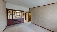This single-floor residence presents a functional and cohesive layout connecting its key living spaces. The living area is finished with low-pile carpet and features textured, tile-patterned wall coverings in warm beige and taupe tones, accented by dark wood crown molding and baseboards throughout. A textured popcorn ceiling adds dimension overhead. A large window near the front entry allows natural light to filter in, offering views of surrounding greenery, while a ceiling fan provides ventilation and additional lighting.

The kitchen is partially separated from the living area by a half-wall pass-through with a dark wood-paneled bar base fitted with open shelving. The kitchen itself is equipped with white upper and lower cabinetry trimmed in dark wood, a granite-patterned laminate countertop, a stainless steel sink, a white electric range, and a full-size refrigerator. Light tile flooring runs throughout the kitchen area.

The bedroom is a generously sized, carpeted room with the same textured wall treatment, a ceiling fan with light fixture, and a window dressed with horizontal blinds providing natural light and outdoor views.

The bathroom features tile-look vinyl flooring, a single vanity with a marble-patterned countertop, a toilet, and a tub-shower combination with a handheld showerhead.