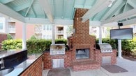 Located in Spanish Fort, this single-level outdoor pavilion is designed for comfortable alfresco gatherings and entertaining. The space is anchored by a striking full-size brick fireplace, which serves as both a functional hearth and a bold architectural centerpiece. Flanking the fireplace are multiple built-in stainless steel grills set into dark countertops, providing ample cooking stations for hosting events of any size. The pavilion is sheltered beneath a vaulted roof with exposed beams painted in a distinctive teal hue, supported by classic white columns that lend the space an inviting, resort-like character. Track lighting is mounted along the beams, ensuring the area remains well-lit during evening gatherings. The open-air design allows for natural light and gentle breezes, while the lush surrounding greenery creates a private, park-like atmosphere. The concrete flooring provides a durable, low-maintenance surface, and additional storage or utility drawers are integrated into the outdoor kitchen build-out. The overall ambiance is relaxed yet well-appointed, blending practical outdoor cooking amenities with warm, classic design elements that make this pavilion a standout communal feature.