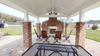 This inviting outdoor pavilion serves as a central gathering space within a residential community, thoughtfully designed for relaxation and social interaction. The structure is anchored by sturdy brick columns that support a crisp white gabled roof with exposed beam detailing, creating a bright and airy atmosphere while providing ample shelter.

At the heart of the pavilion stands a substantial brick fireplace, serving as a natural focal point and gathering spot. Surrounding it, a set of wicker-style swivel lounge chairs arranged on a decorative paver patio offers a comfortable seating area ideal for conversation and leisure. Additional metal mesh outdoor tables and seating are also present throughout the covered space.

The paver patio extends beyond the covered structure, connecting the pavilion to the broader community amenities. Views from the pavilion encompass a fenced swimming pool, a playground area with colorful equipment, and well-maintained green lawns. Outdoor grills are accessible on the open patio, making the area well-suited for outdoor dining and entertaining.

The surrounding community features multi-story residential buildings, mature trees, and landscaped grounds visible from all sides of the pavilion, giving the space a connected, neighborhood-oriented feel. Overall, this pavilion offers a well-equipped and attractive outdoor amenity space designed to serve the needs of an active residential community.
