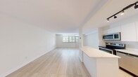 This well-appointed 1-bedroom, 1-bathroom unit offers a thoughtfully designed open-concept layout that maximizes the efficient use of space. Light wood-toned flooring flows seamlessly throughout, lending a clean and contemporary feel to every room. The combined kitchen and living area forms the heart of the unit, offering a generous, airy feel. The modern kitchen is outfitted with sleek, high-gloss white cabinetry extending to the ceiling, stainless steel appliances including a refrigerator, range, microwave, and dishwasher, a white subway tile backsplash, and a marble-topped peninsula that doubles as a breakfast bar. Track lighting overhead adds a stylish, functional touch. The open living area is bathed in natural light from a large picture window framing views of lush greenery outside. The bedroom is a comfortable, well-proportioned retreat with its own window offering garden views and ample natural light. A convenient in-unit stacked washer and dryer is tucked neatly behind a sliding closet door. The bathroom features a full soaking tub with an overhead shower, framed by crisp white subway tile. The unit's neutral palette of whites and soft beiges creates a bright, inviting ambiance throughout.